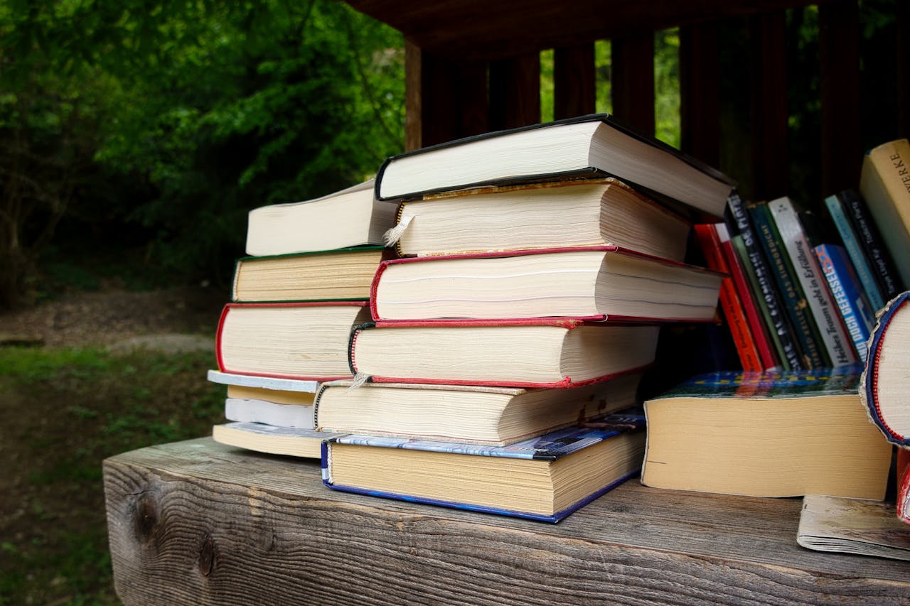 A pile of assorted books on a rustic wooden table in a lush outdoor setting.